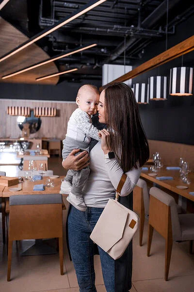 Eine stilvolle Mutter mit Baby im Café; die hellcremefarbene Älly Cappuccino Kompakt-Wickeltasche hängt elegant am Handgelenk.