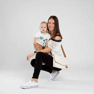 Studio-Lifestyle-Foto einer lächelnden Mutter mit Baby, die die hellcremefarbene Älly Cappuccino Wickeltasche aus veganem Leder bequem quer über dem Körper trägt.