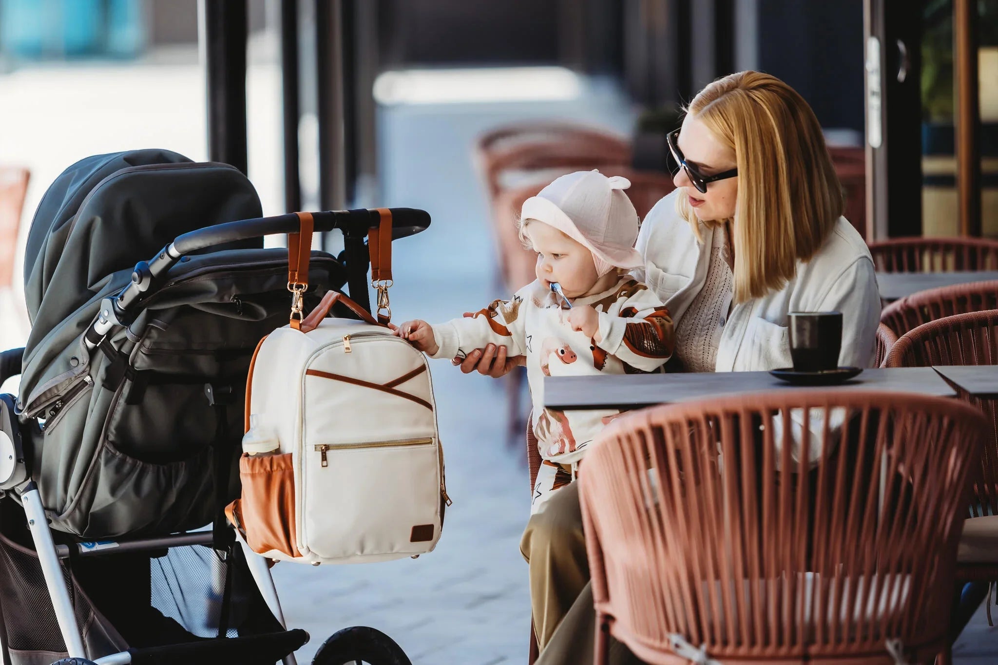 Mamma e bambino in un bar con lo zaino fasciatoio Ally Scandic (cappuccino) comodamente appeso al manubrio del passeggino tramite le cinghie in dotazione.