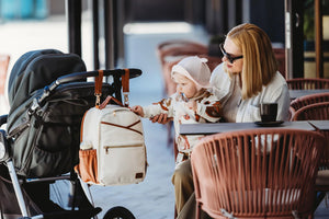 Mamma e bambino in un bar con lo zaino fasciatoio Ally Scandic (cappuccino) comodamente appeso al manubrio del passeggino tramite le cinghie in dotazione.