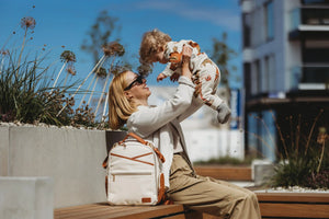 Mamma che solleva giocosamente il suo bambino all'aperto con lo zaino fasciatoio Ally Scandic (bianco e cappuccino) appoggiato sulla panchina accanto a lei.