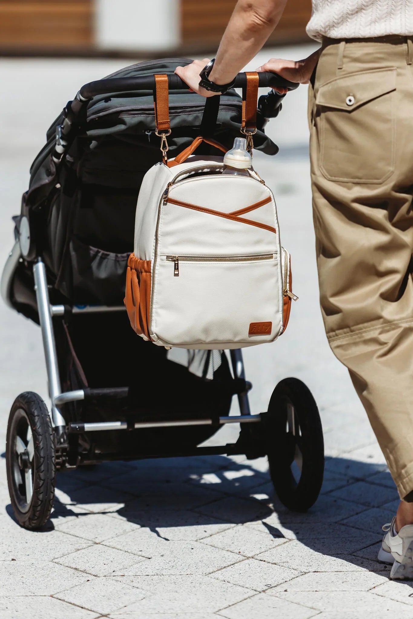 Genitore che spinge un passeggino all'aperto con lo zaino fasciatoio Ally Scandic color cappuccino appeso in sicurezza al manubrio.
