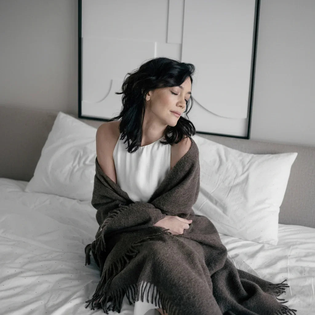 Femme aux yeux fermés, l'air calme et détendu, enveloppée dans un jeté en laine marron foncé à franges sur un lit aux draps blancs dans une chambre moderne.