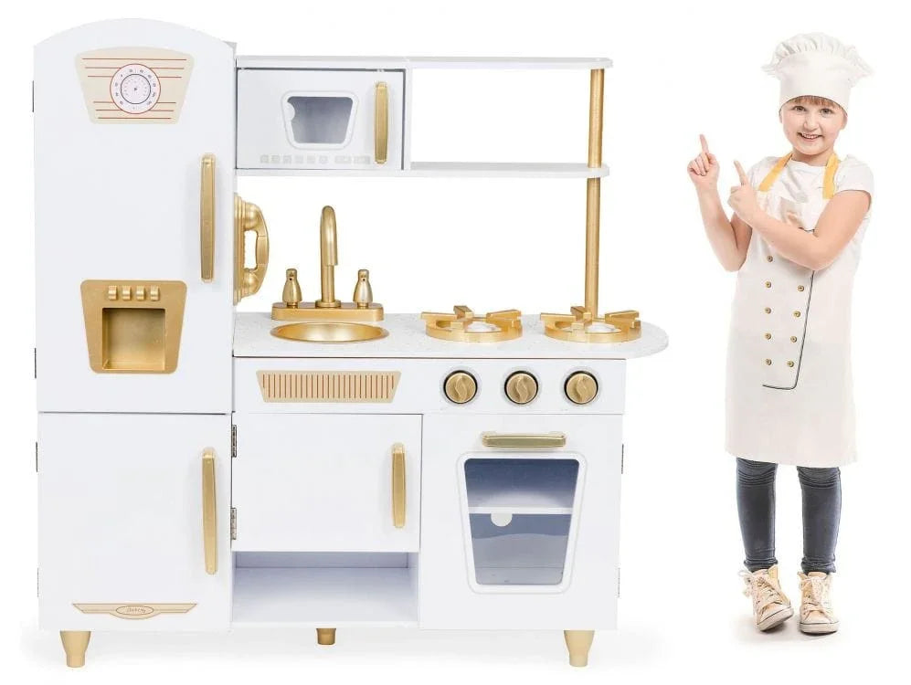 Jeune fille souriante en tablier et toque de chef pointant avec enthousiasme la grande cuisine jouet en bois Angelove blanche et dorée.