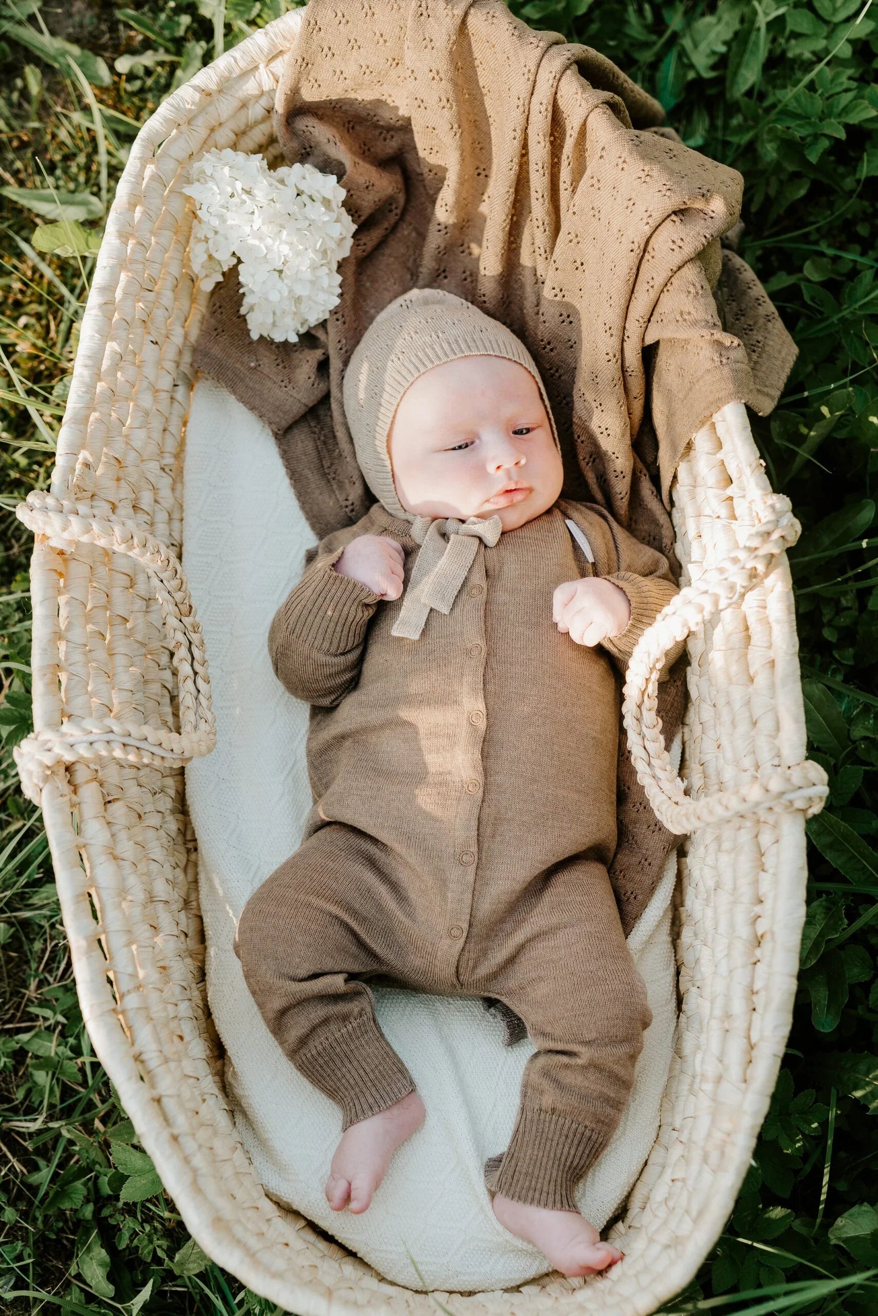 Baby im braunen Strickstrampler und Haube, liegt in einem Weidenkorb auf grünem Gras