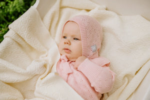 Bébé fille avec bonnet et pull en tricot rose sur une couverture texturée crème dans son berceau