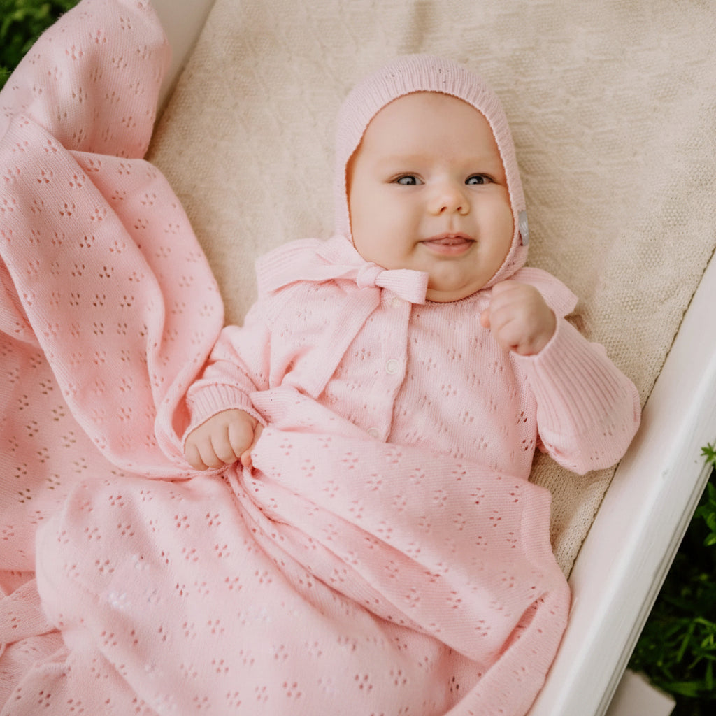 baby girl in pink knitted romper and bonnet with matching blanket in cradle
