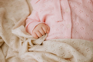 Baby hand holding cream knit blanket, wearing pink knitwear with delicate pattern