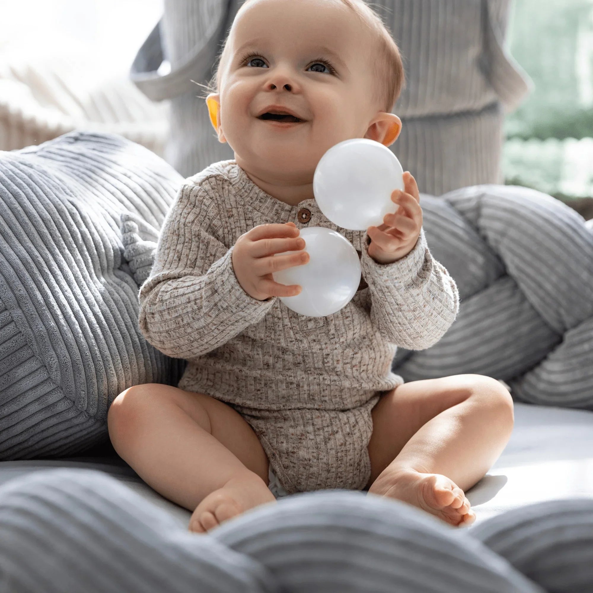 Baby sitzt in einem grauen, geflochtenen Nestchen-Spielkissen, hält weiße Bälle, texturierter Stoff