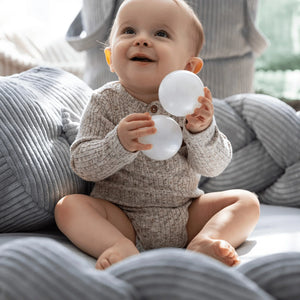 Baby sitzt in einem grauen, geflochtenen Nestchen-Spielkissen, hält weiße Bälle, texturierter Stoff