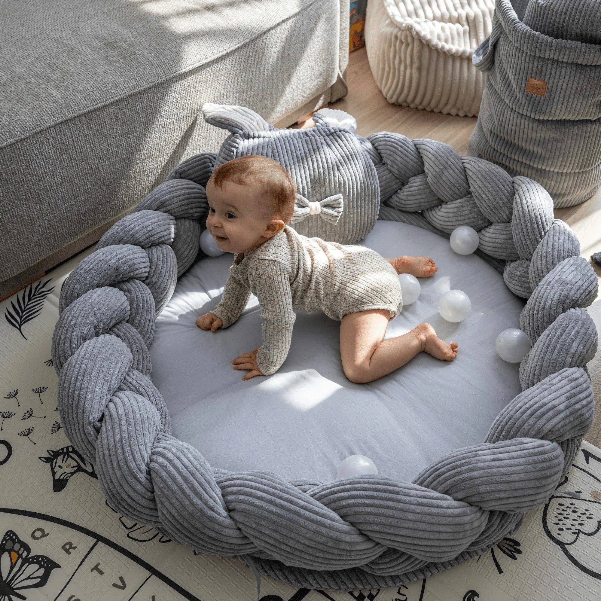 Baby im runden, grauen Nestchen-Spielkissen mit weicher Umrandung und Plüschstoff