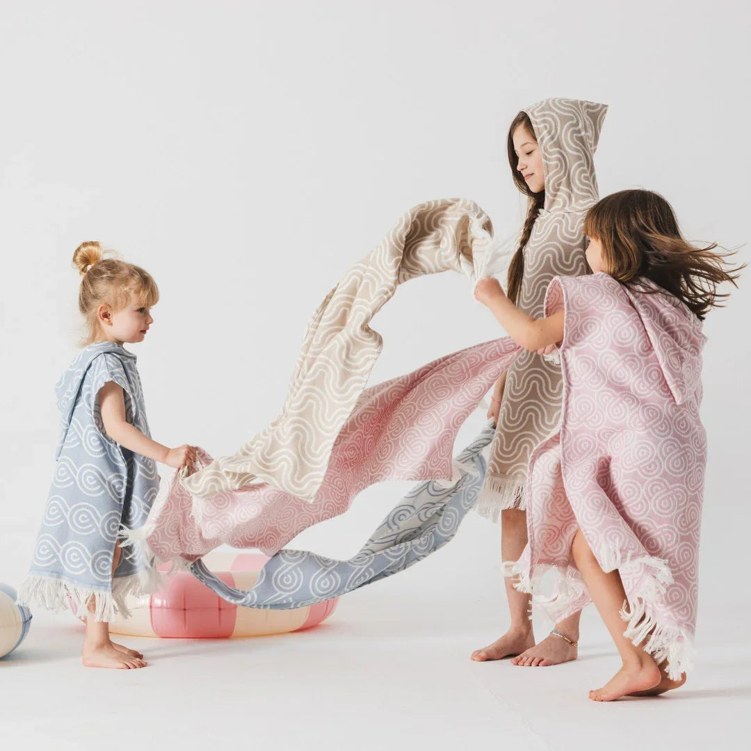 Kids wearing hooded bamboo bath ponchos in sand, pink, and blue swirl patterns with fringe trim