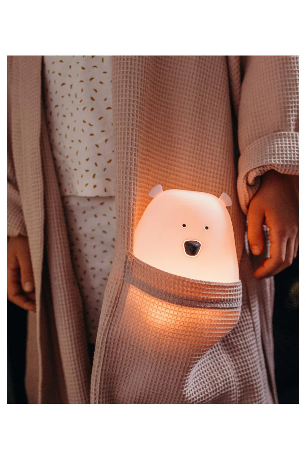 Veilleuse ourson rose avec télécommande dans la poche d'un peignoir gaufré, diffusant une lueur chaleureuse