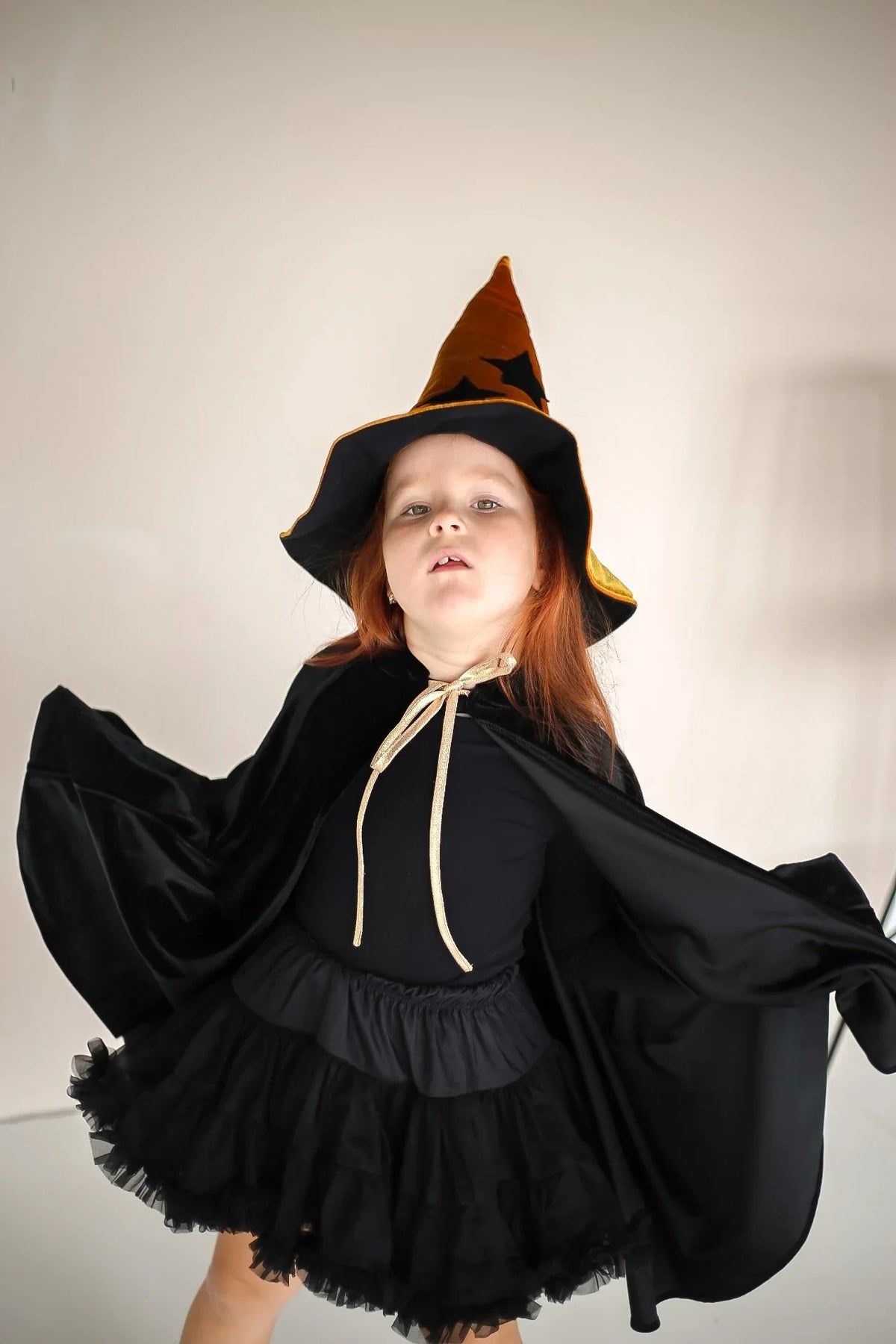 Child posing in black velour cape, tutu and orange-black witch hat