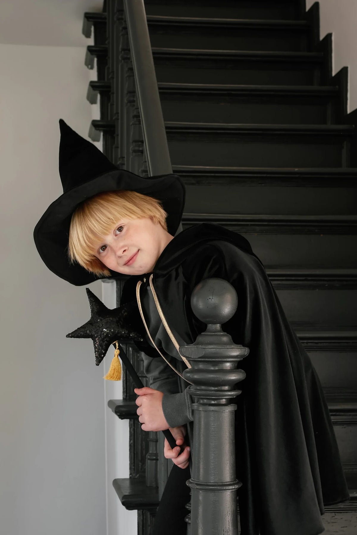 Child in black velour cape and witch hat holding black star wand beside staircase