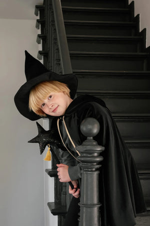 Child in black velour cape and witch hat holding black star wand beside staircase