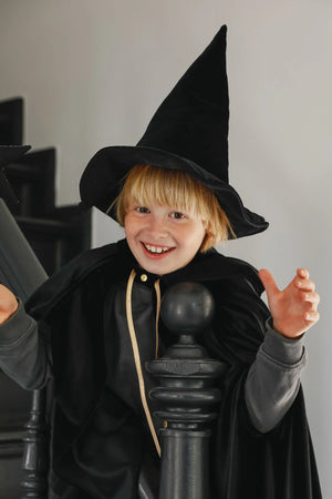 Smiling child in black Moi Mili cape and pointed witch hat on staircase