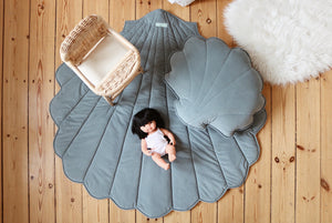 Blue-grey Moi Mili shell playmat styled with a doll, rattan chair and matching shell cushion on wooden flooring