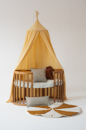 Butter yellow Moi Mili cotton canopy styled above a round wooden children’s bed with striped cushions and a flower-shaped play mat