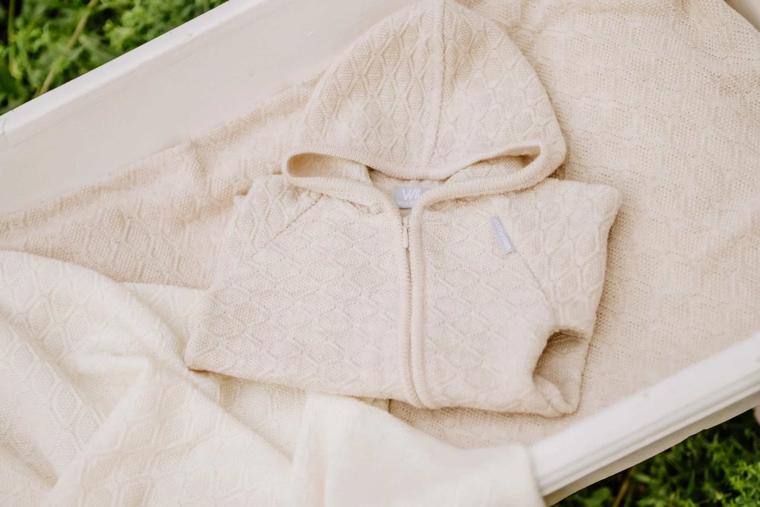 Grenouillère à capuche en tricot crème avec motif losanges sur une couverture assortie dans un berceau