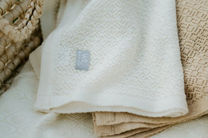 Couverture bébé ajourée en tricot crème, motif losanges, présentée dans un panier en osier