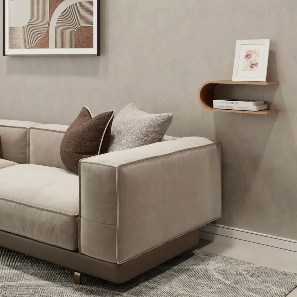 Sofá de terciopelo beige con cojines marrones y grises, estante de pared moderno y arte abstracto en una sala neutra.