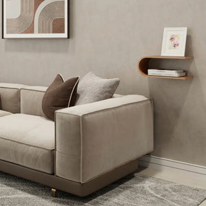 Sofá de terciopelo beige con cojines marrones y grises, estante de pared moderno y arte abstracto en una sala neutra.