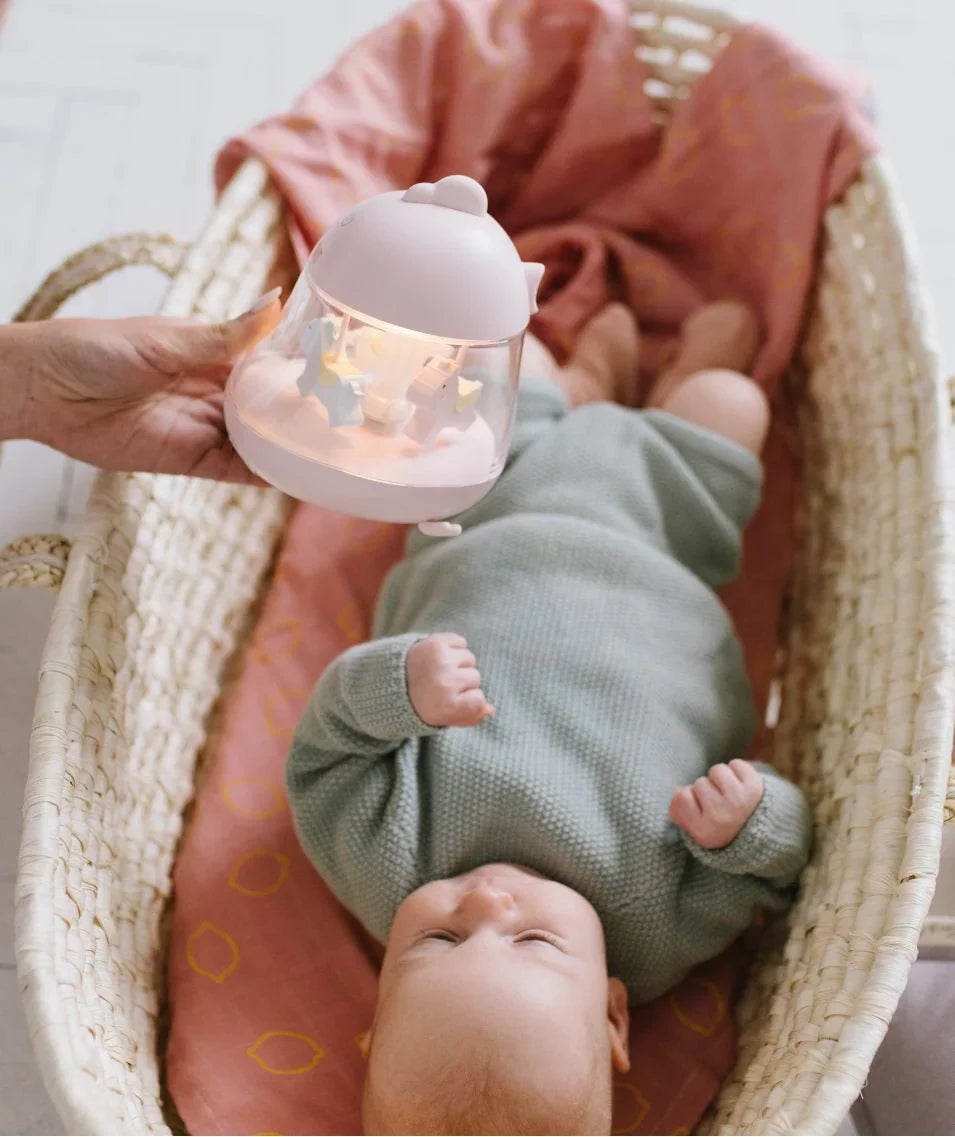 Bébé allongé dans un couffin en osier avec couverture rose, adulte tenant une veilleuse pastel avec figurines rotatives