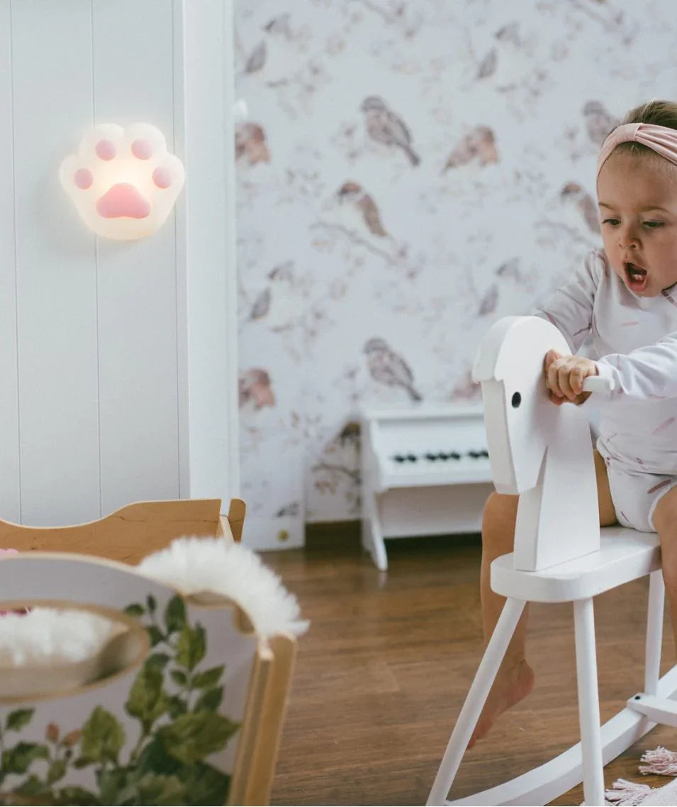 Nursery room with child on white rocking horse, pink paw night light on wall, bird wallpaper, and floral toy basket