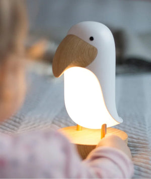 Luz nocturna infantil en forma de pájaro con cuerpo blanco, pico y base de madera, sobre una cama