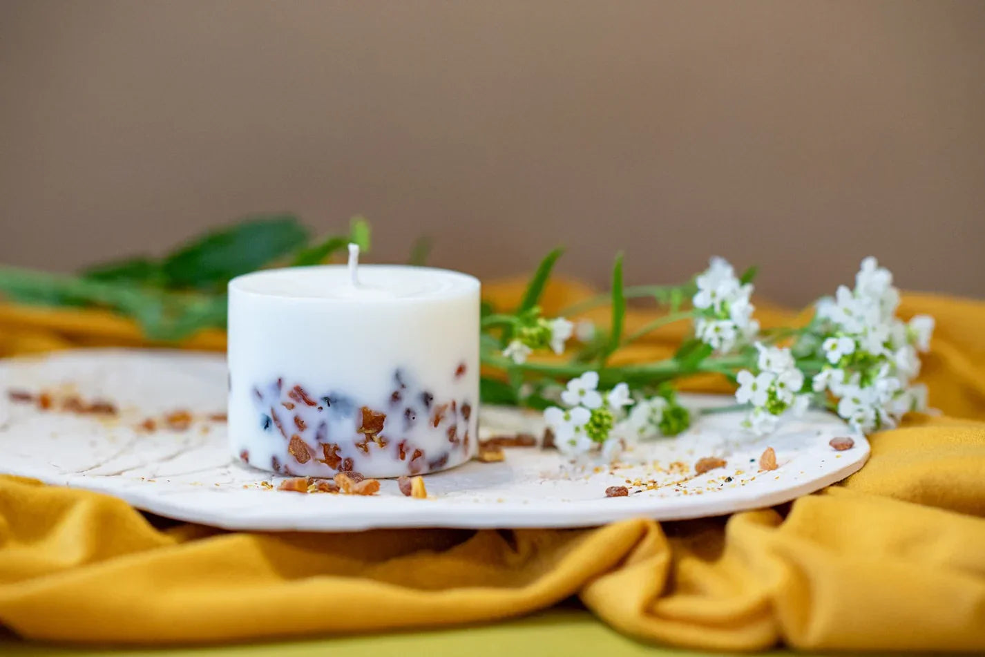 Candela in cera di soia artigianale con inclusioni botaniche su un piatto in ceramica, circondata da fiori bianchi