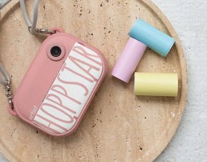 Fotocamera per bambini Hoppstar in rosa con tre rotoli di carta colorata: blu, rosa e giallo, su un vassoio rotondo