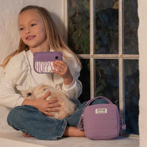 Bambino con fotocamera giocattolo Hoppstar color lavanda, borsa abbinata, coniglietto morbido, su seduta casual accanto alla finestra