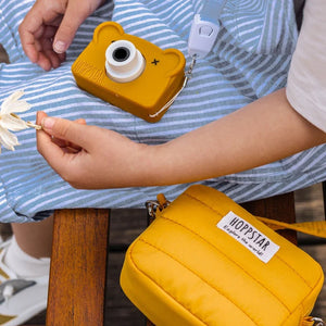 Mini borsa fotografica Hoppstar gialla e fotocamera per bambini a forma di orsetto con tracolla in tessuto sulle ginocchia di un bambino