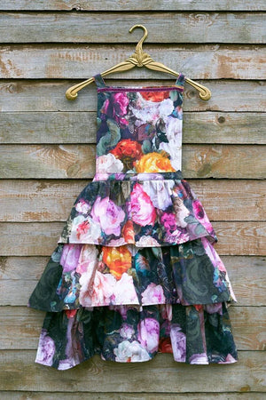 Floral tiered dress with pink, orange, and purple roses on dark background, hanging on gold hanger against wood wall