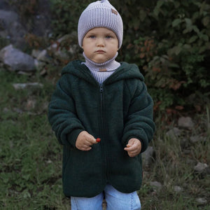 Toddler in forest green fleece hoodie, light gray knit beanie, and turtleneck, holding red berry outdoors