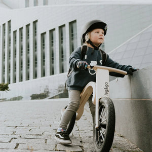 Child riding white leg&go wooden balance bike with black tires on cobblestone path