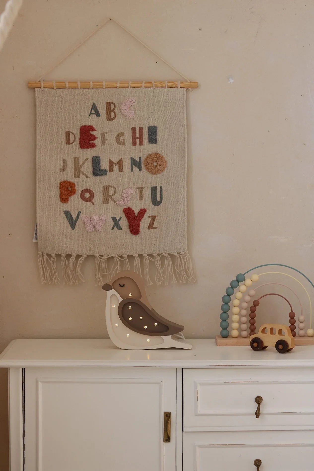 Handwoven nursery alphabet wall hanging with colorful felt letters, wooden rod, and tassels, displayed above a white dresser with wooden toys.