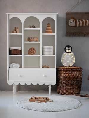 White nursery bookshelf with arched compartments, scalloped base, and drawer, styled with wooden toys and wicker baskets