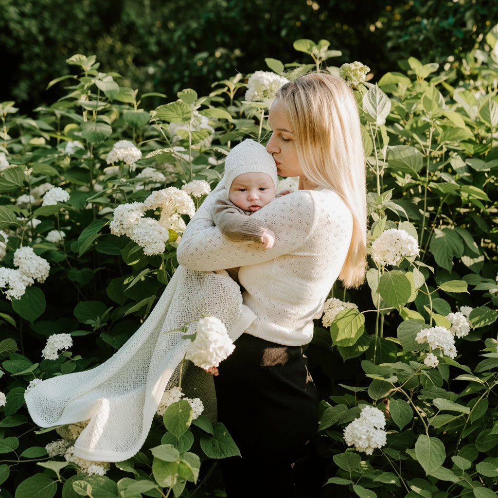 Maman tenant son bébé enveloppé dans une couverture crème en tricot parmi des hortensias blancs dans un jardin