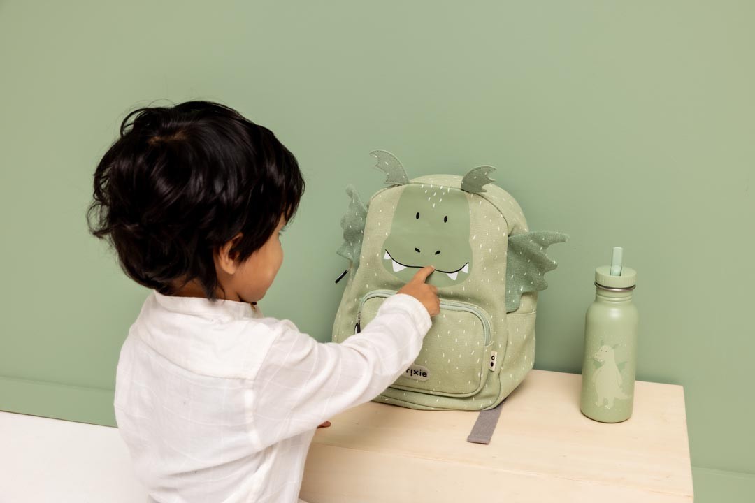 Child touching the face of a Trixie Mr. Dragon backpack beside a matching green water bottle