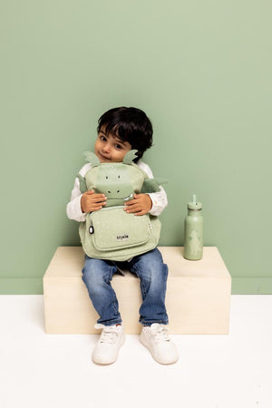 Child sitting and hugging the green Trixie Mr. Dragon backpack beside a matching bottle