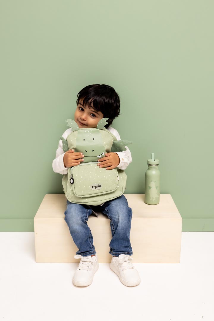 Child sitting and hugging the green Trixie Mr. Dragon backpack beside a matching bottle
