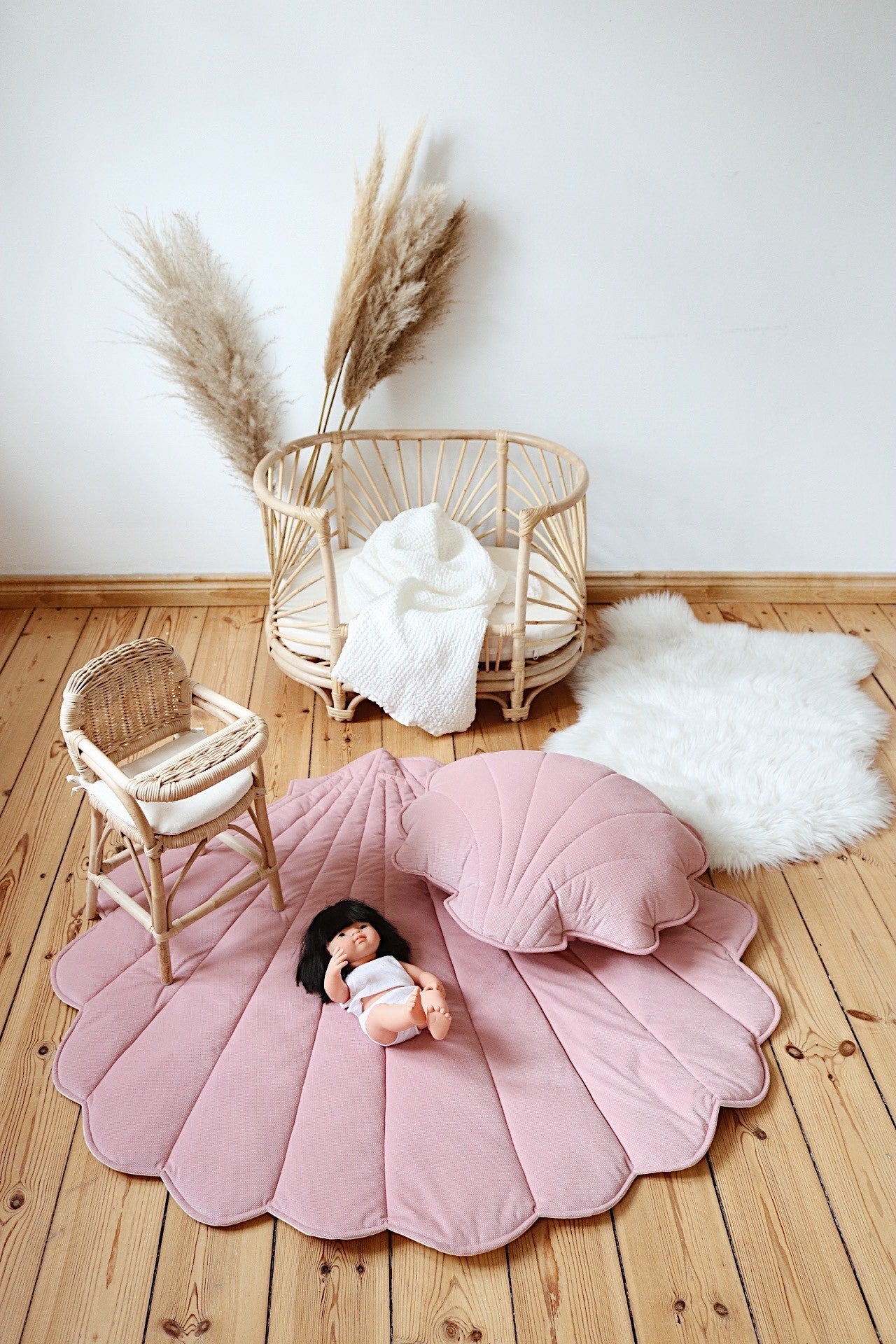 Doll resting on a powder pink shell playmat with a matching cushion and rattan nursery furniture