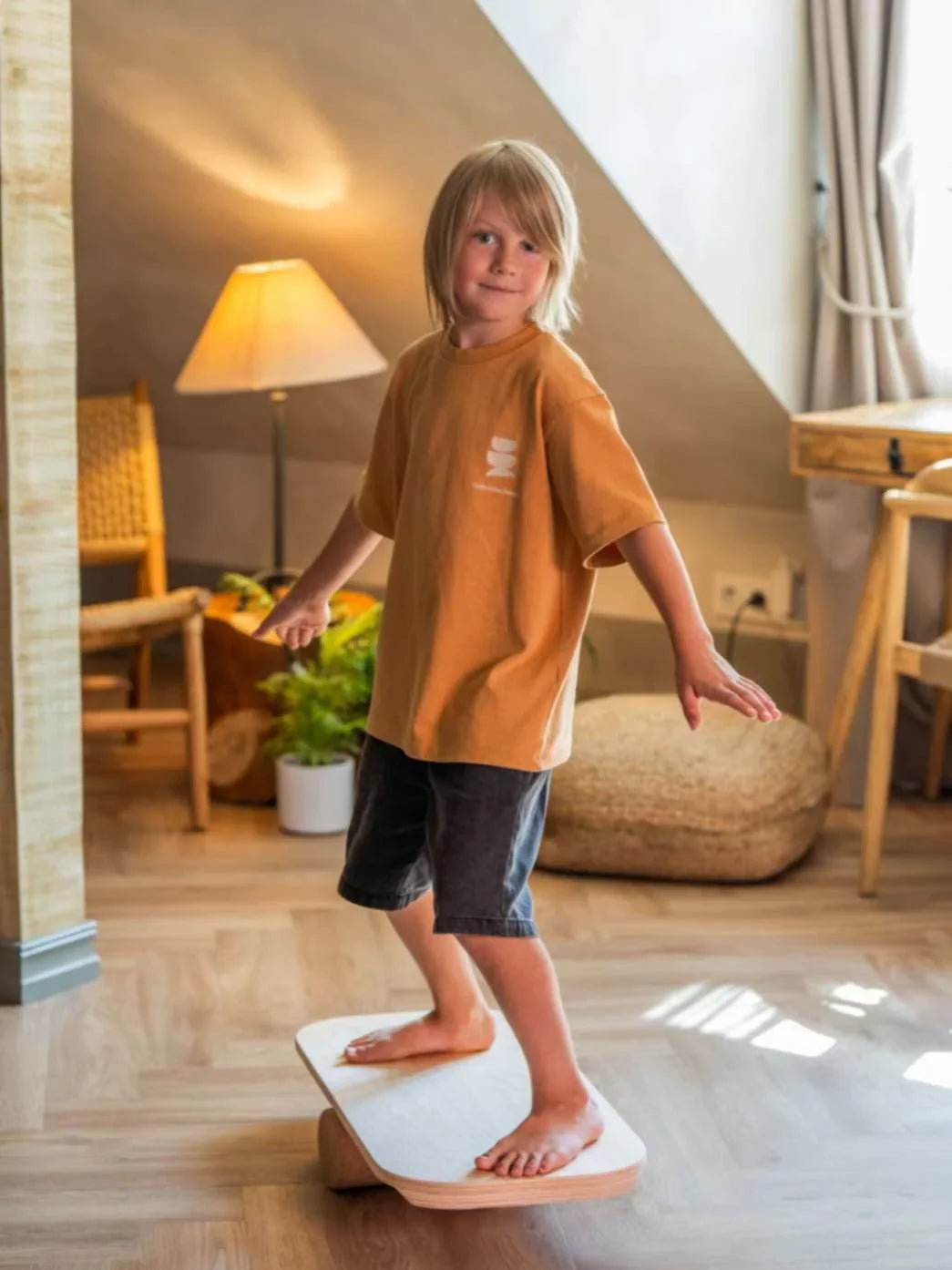Niño manteniendo el equilibrio sobre una tabla de madera rectangular con rodillo de ejercicio en interiores