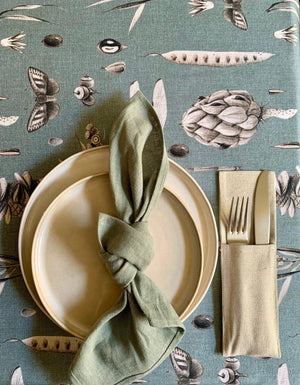 Sage green linen napkin tied in knot on ceramic plates with botanical tablecloth and cutlery holder