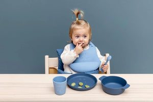 Tout-petit avec un set bol, assiette, gobelet et bavoir en silicone bleu sur table en bois clair