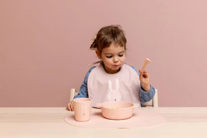 Enfant utilisant un bol et un gobelet en silicone rose avec motif lapin, avec bavoir et set de table assortis sur la table