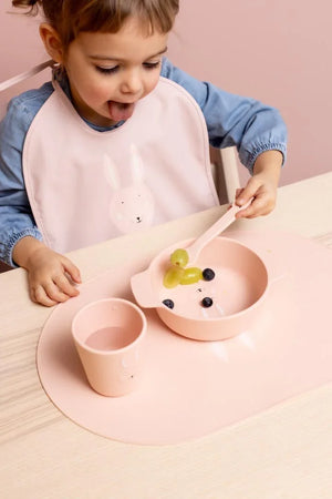 Tout-petit mangeant avec un bol, un gobelet et une cuillère en silicone rose poudré arborant un mignon motif tête de lapin