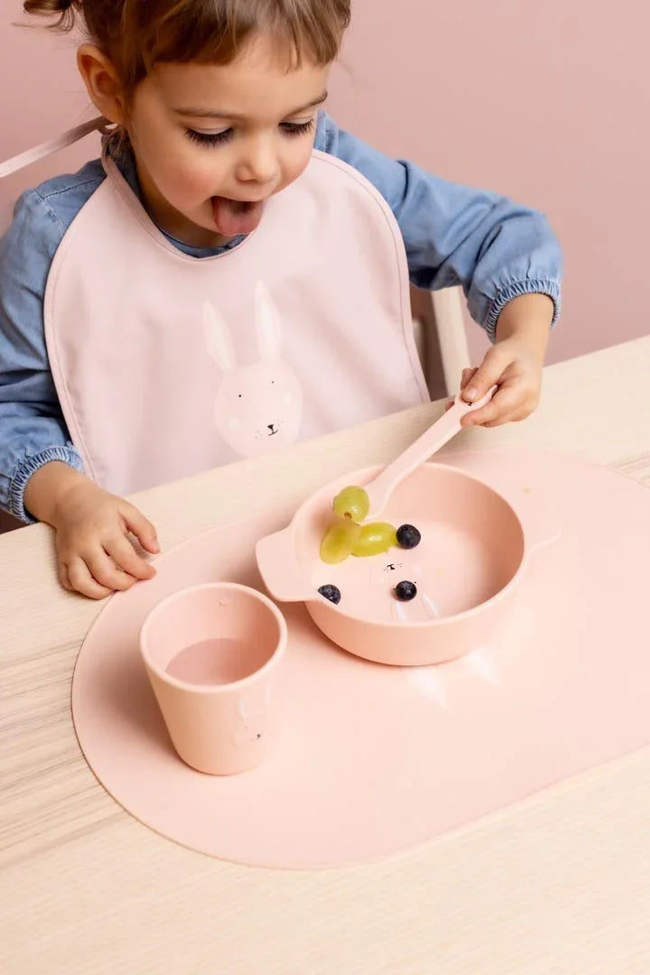 Niño pequeño comiendo con bol, vaso y cuchara de silicona rosa con un tierno diseño de cara de conejo